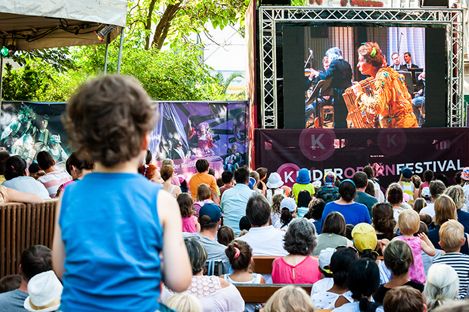 Foto: Kinderopern Festival © stadt wien marketing / Johannes Wiedl Photo with a screen showing an opera performance, in front of it the audience consisting mainly of children