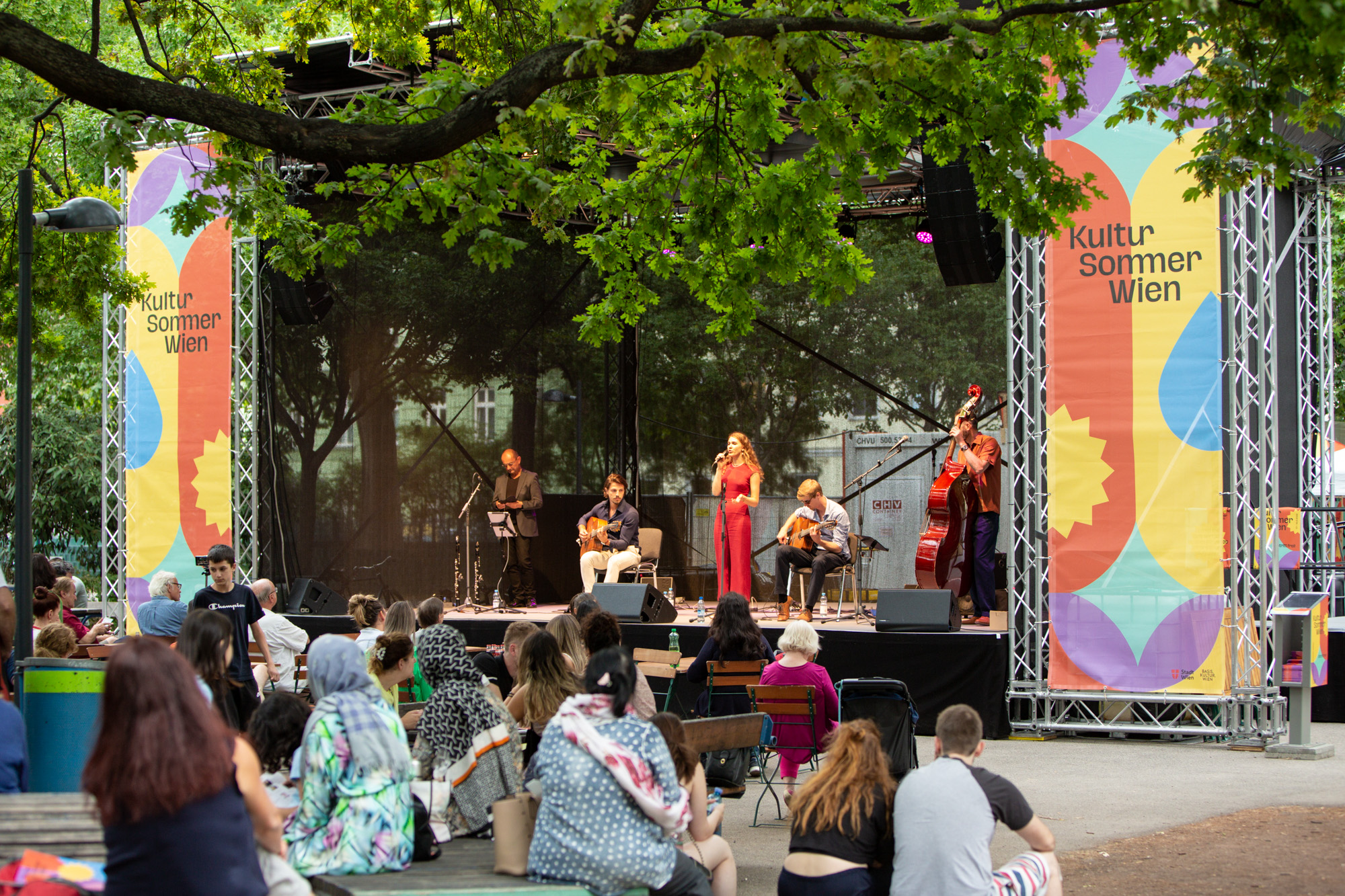 Kultursommer Wien / Mortarapark, Belle Affaire © Judith Stehlik Foto einer Open Air-Situation in einem Park: eine Band musiziert auf einer Bühne unter Bäumen, davor das Publikum