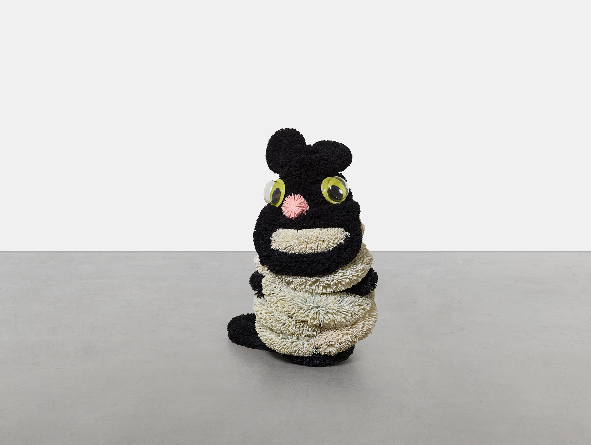 © Damien Hirst and Science Ltd. All rights reserved/ Bildrecht, Wien 2025 Das Foto zeigt eine von Damien Hirst geschaffene Skulptur einer sitzenden Katze aus flauschigem schwarzem und beigen Material mit großen grüngelben Plaktikaugen
