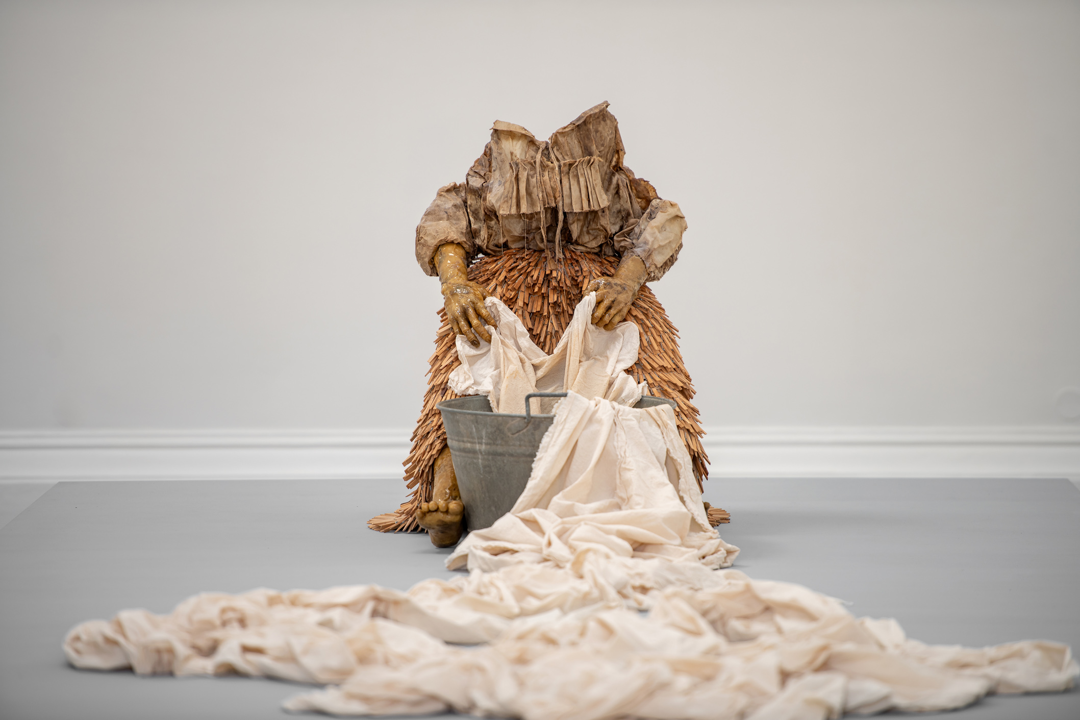 Shannon Alonzo. Washerwoman im Theseustempel - Ausstellungsansicht © KHM-Museumsverband / Foto: Thomas Ritter Das Foto zeigt eine Mixed-Media-Skluptur, eine lebensgroße gesichtslose Figur aus Harz, Bienenwachs, Wäscheklammern, brauner Baumwolle und einem mit Wasser gefüllten Waschtrog