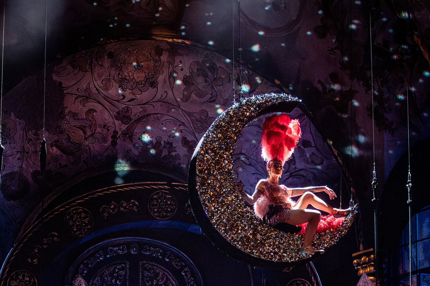 Wiener Staatsoper / Tannhäuser © Foto: Ashley Taylor Das Szenefoto aus der Oper "Tannhäuser" zeigt einen glitzernden Mond, der über der Bühne hängt. Darauf sitzt eine der Opernsängerinnen gekleidet wie eine Revuetänzerin mit großem rotem Kopfschmuck. Die Szene wird von hinten beleuchtet.