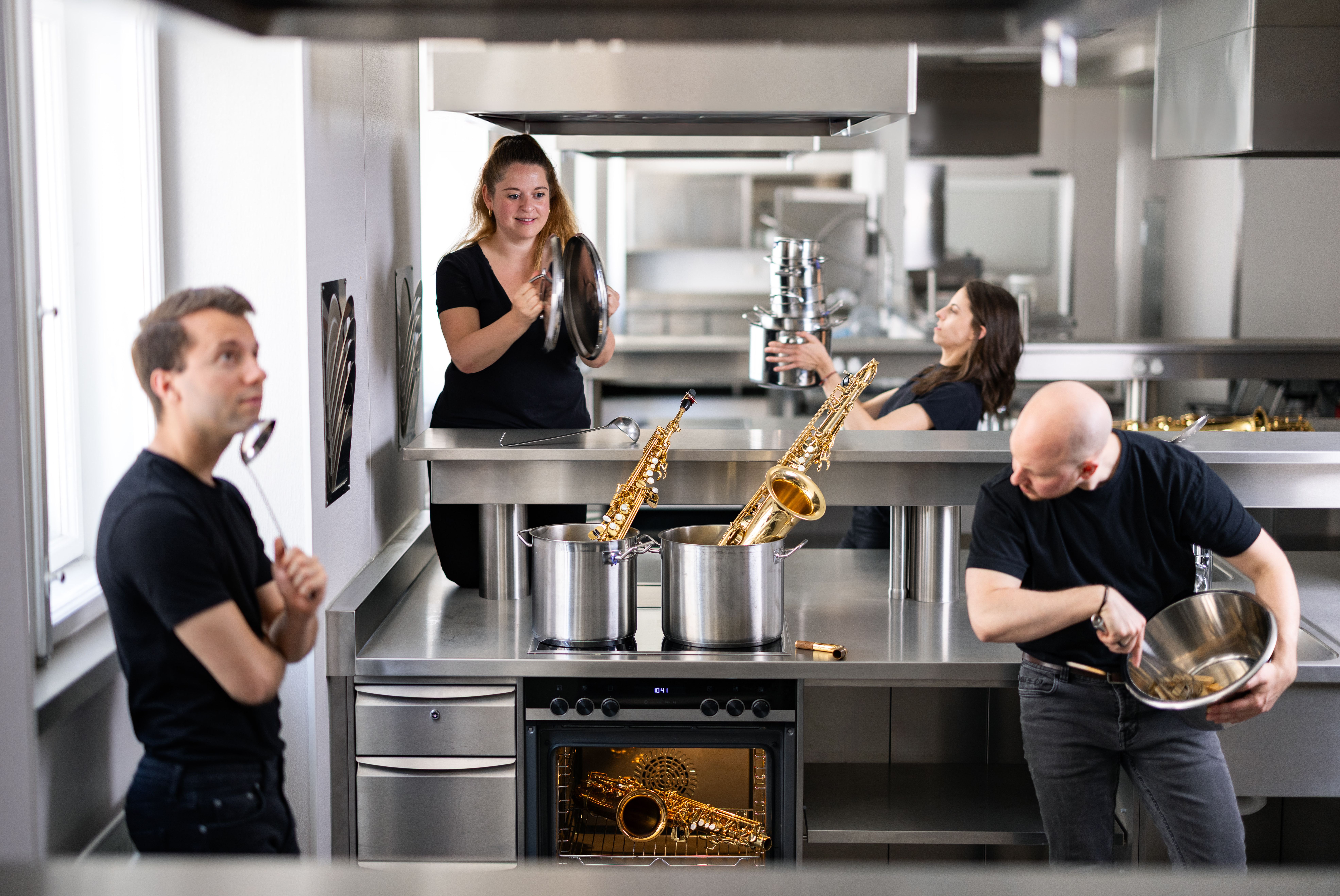 Aureum Saxophon Quartett © Sofija Palurovic The photo shows the Aureum Saxophone Quartet in the kitchen of a restaurant. The four musicians: dressed in black, with their saxophones between stainless steel pots and kitchen surfaces