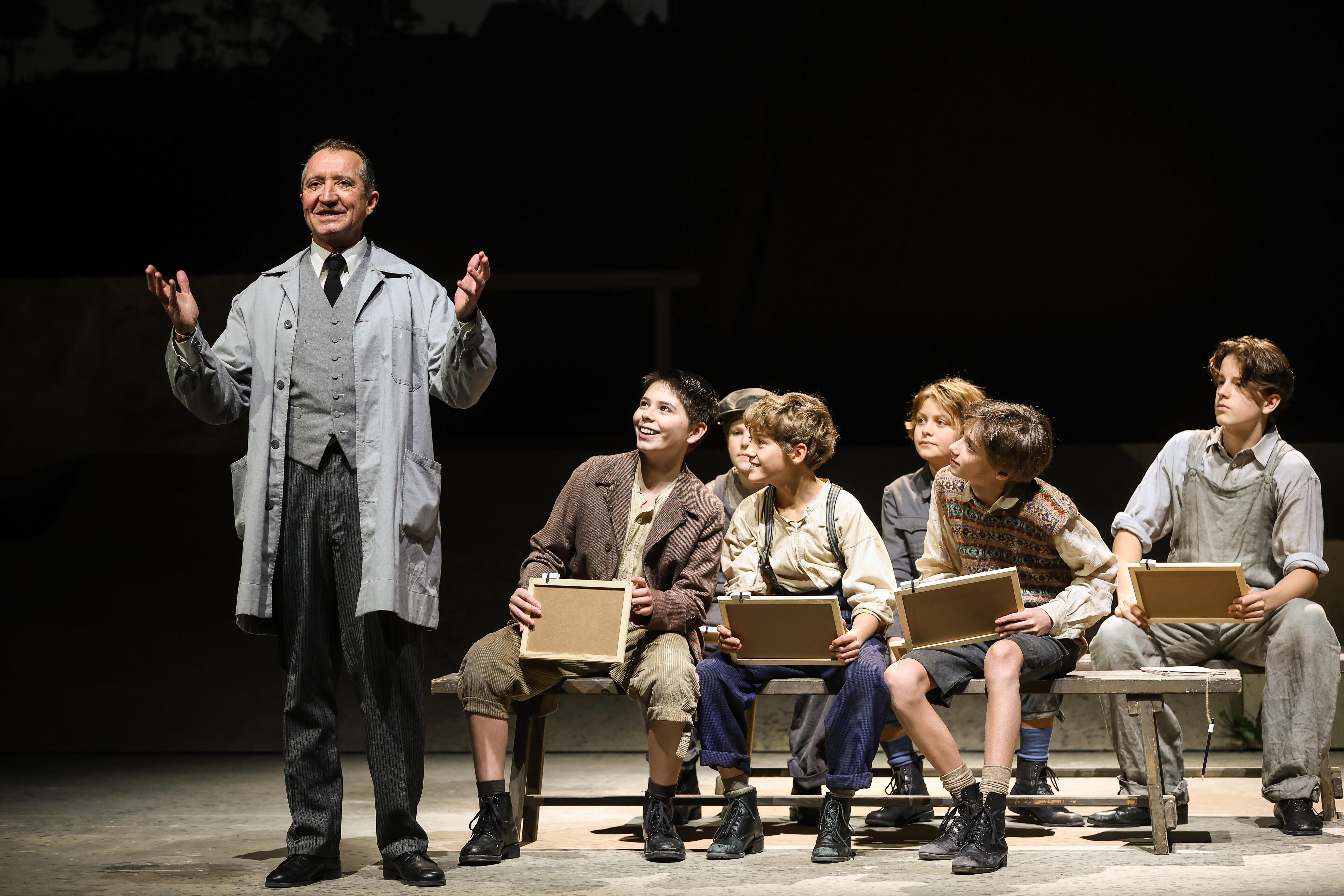 Volksoper Wien / Der Krieg der Knöpfe: Nicolaus Hagg (Louis, der Lehrer), Kinderchor der Volksoper Wien © Barbara Pálffy / Volksoper Wien Das Szenefoto aus dem Stück Der Krieg der Knöpfe zeigt einen Lehrer in grauem Arbeitsmantel, daneben eine Gruppe von Schülern in historischen Kostümen mit Schiefertafeln auf zwei Bänken sitzend