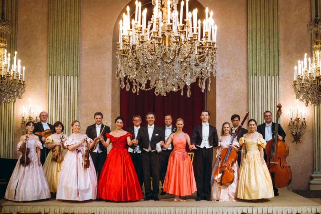 Wiener Residenzorchester © Philipp-Lipiarski Foto des Wiener Residenzorchesters, die Mitglieder in festlicher Abendrobe und mit Instrumenten, in einem historischen Saal mit großen Kronleuchtern