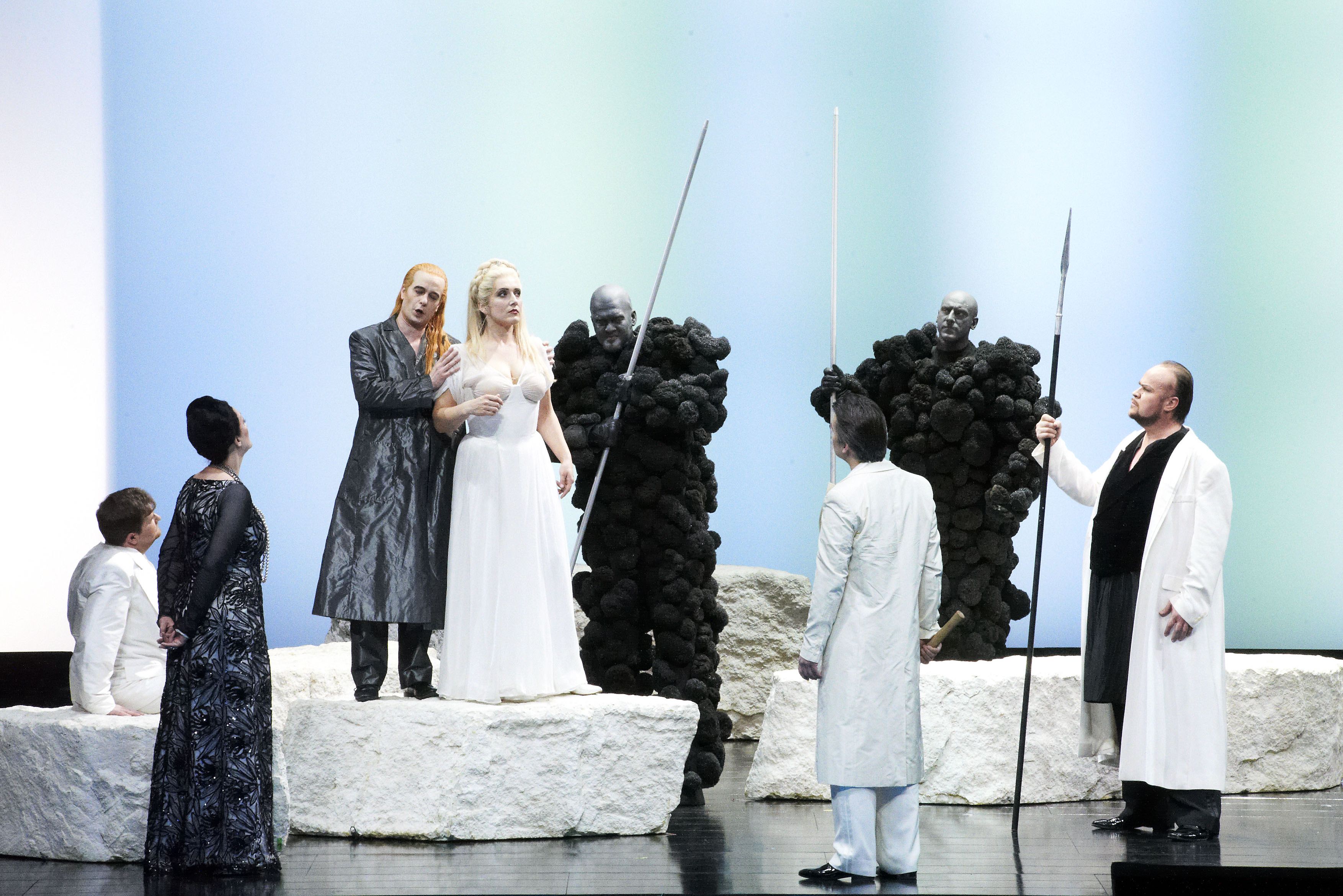 Wiener Staatsoper / Das Rheingold © Foto: Michael Poehn The scene photo from the opera “Das Rheingold” from the Ring of the Nibelungs shows the singers in black and white costumes on a light-colored stage. In the center is a blonde woman in a white dress, next to her a man with long red hair in a black coat