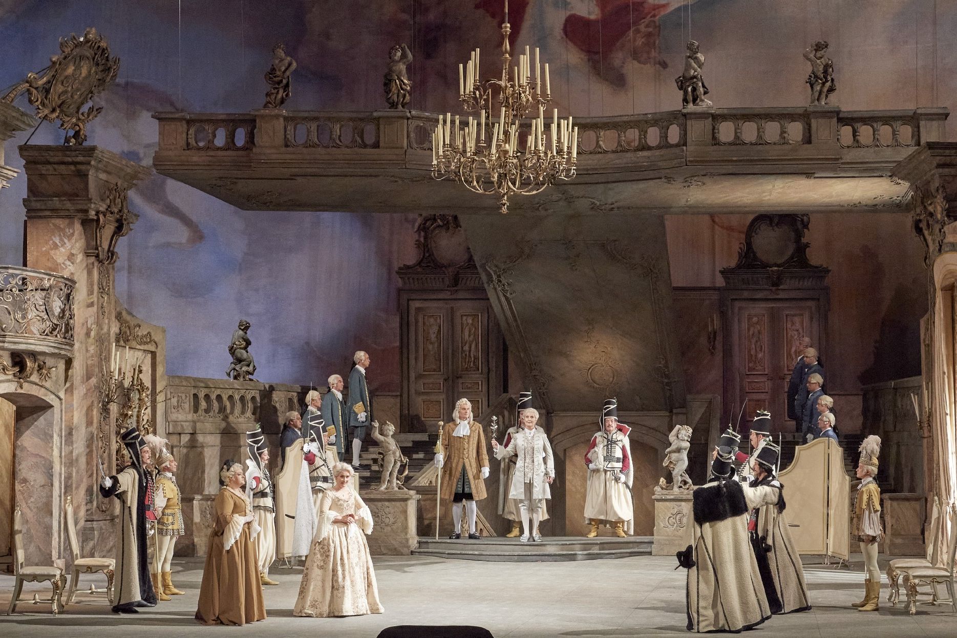Wiener Staatsoper / Der Rosenkavalier © Foto: Michael Poehn The scene photo from the opera “Der Rosenkavalier” of the Vienna State Opera shows a baroque hall in which numerous members of a court are located. The historical costumes and the hall are in shades of beige and brown.