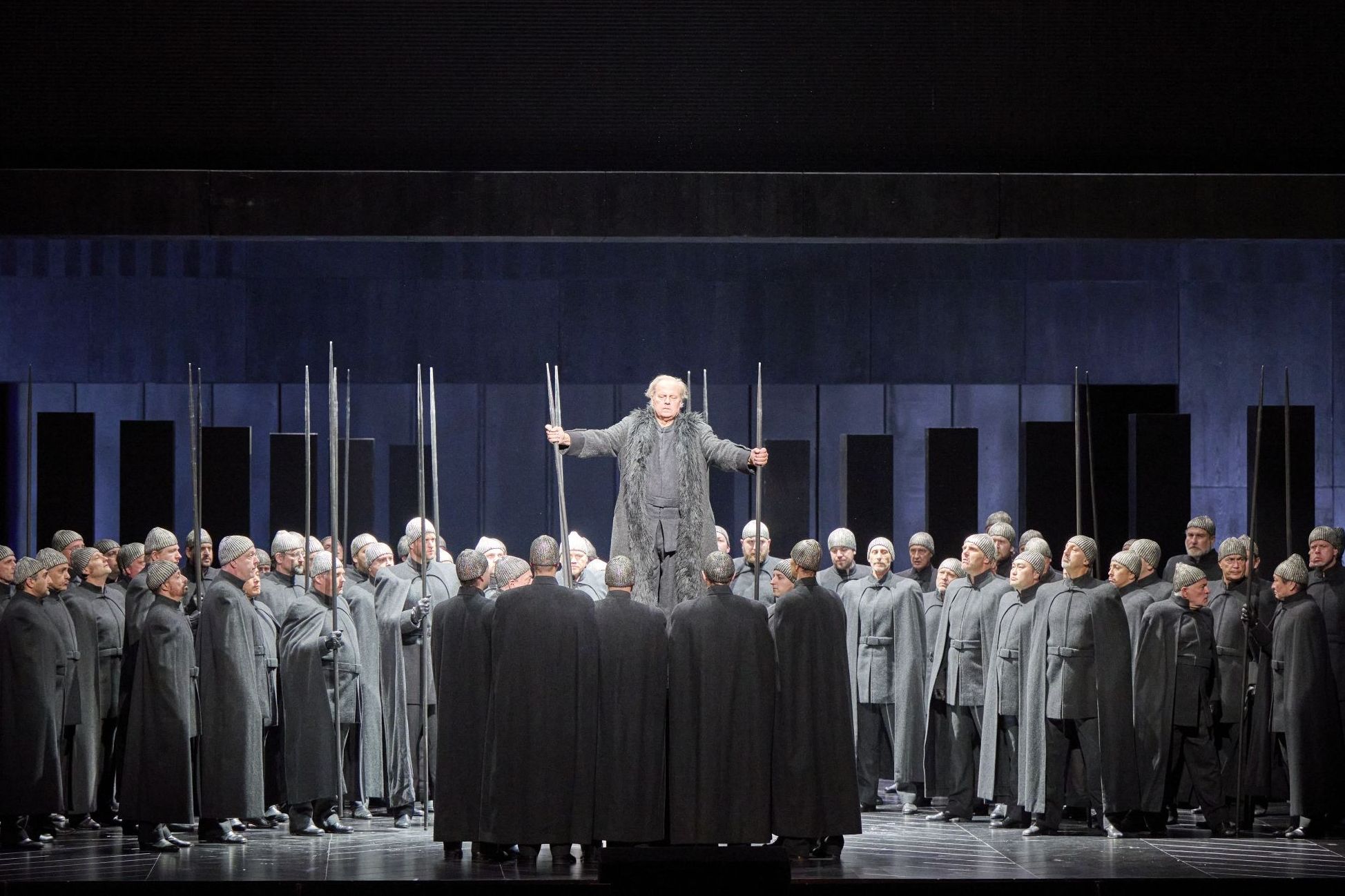 Wiener Staatsoper / Götterdämmerung © Foto: Michael Poehn The scene photo from the opera “Götterdämmerung” from the Ring of the Nibelungs shows a crowd of people in gray uniform-like costumes, in the middle the main actor standing elevated above this crowd, all in front of a dark background