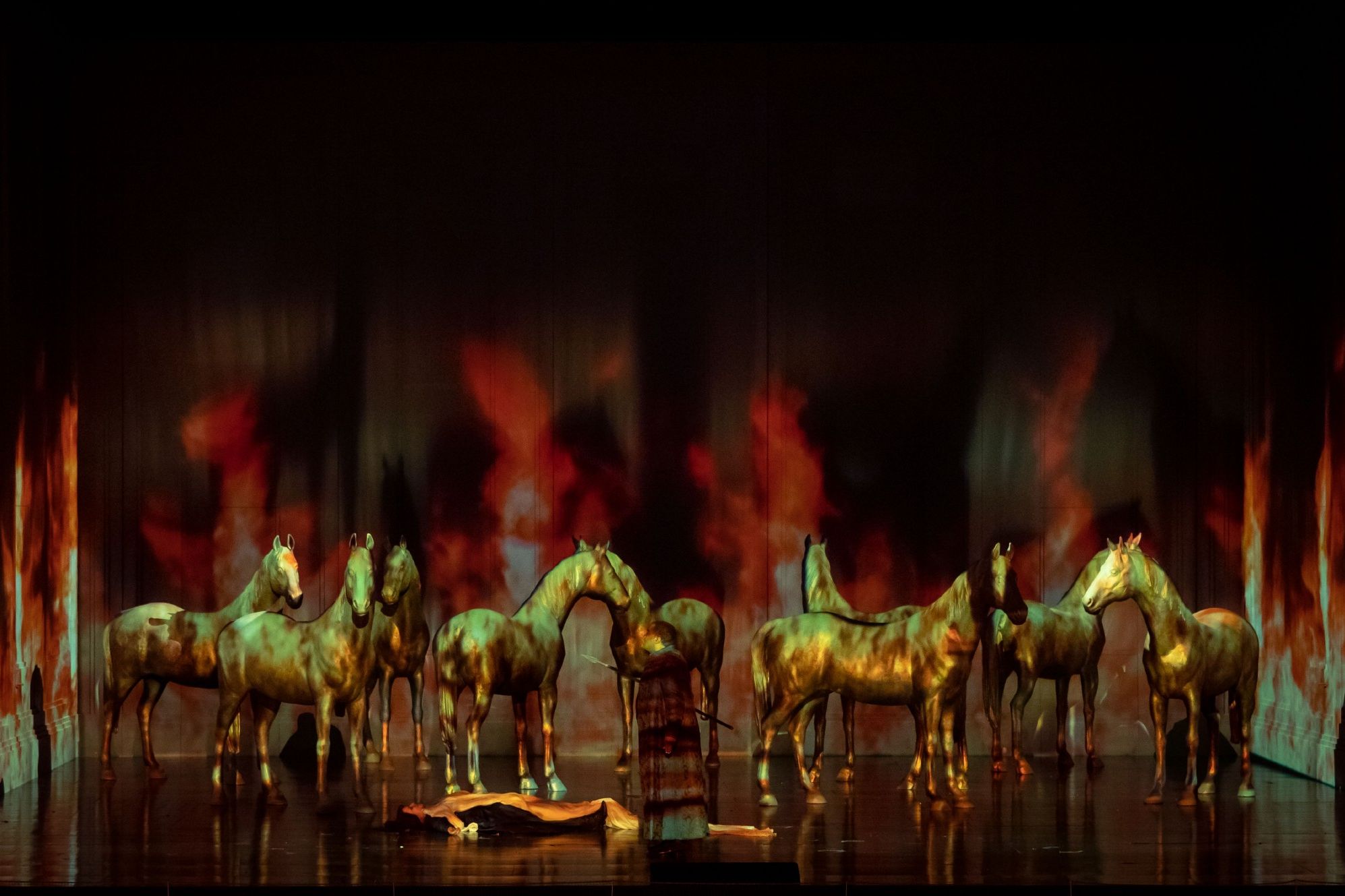 Wiener Staatsoper / Walküre © Foto: Ashley Taylor Scene from the opera “Walküre” at the Vienna State Opera. In the photo, a person is lying on the stage floor, a person with a spear in their hand is standing next to them, behind them golden horses against a black and red background