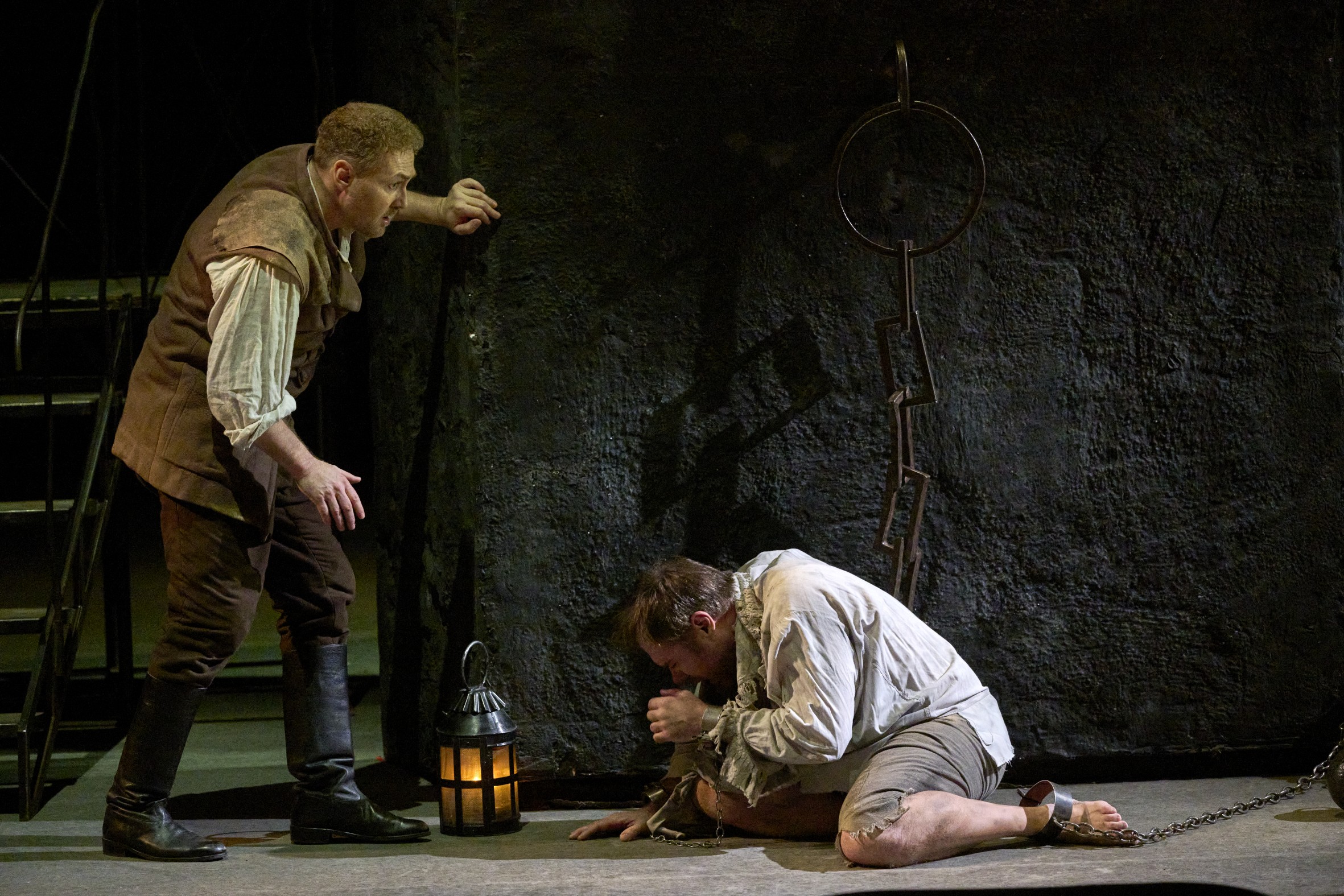 Wiener Staatsoper / Fidelio @ Foto: Michael Poehn Das Szenefoto aus der Oper "Fidelio" zeigt zwei Männer in einem Verlies, einer kniet in zerlumptem Kostüm angekettet am Boden und wendet den Kopf nach untern, der andere stützt sich an der Wand, zwischen ihnen eine brennende Laterne