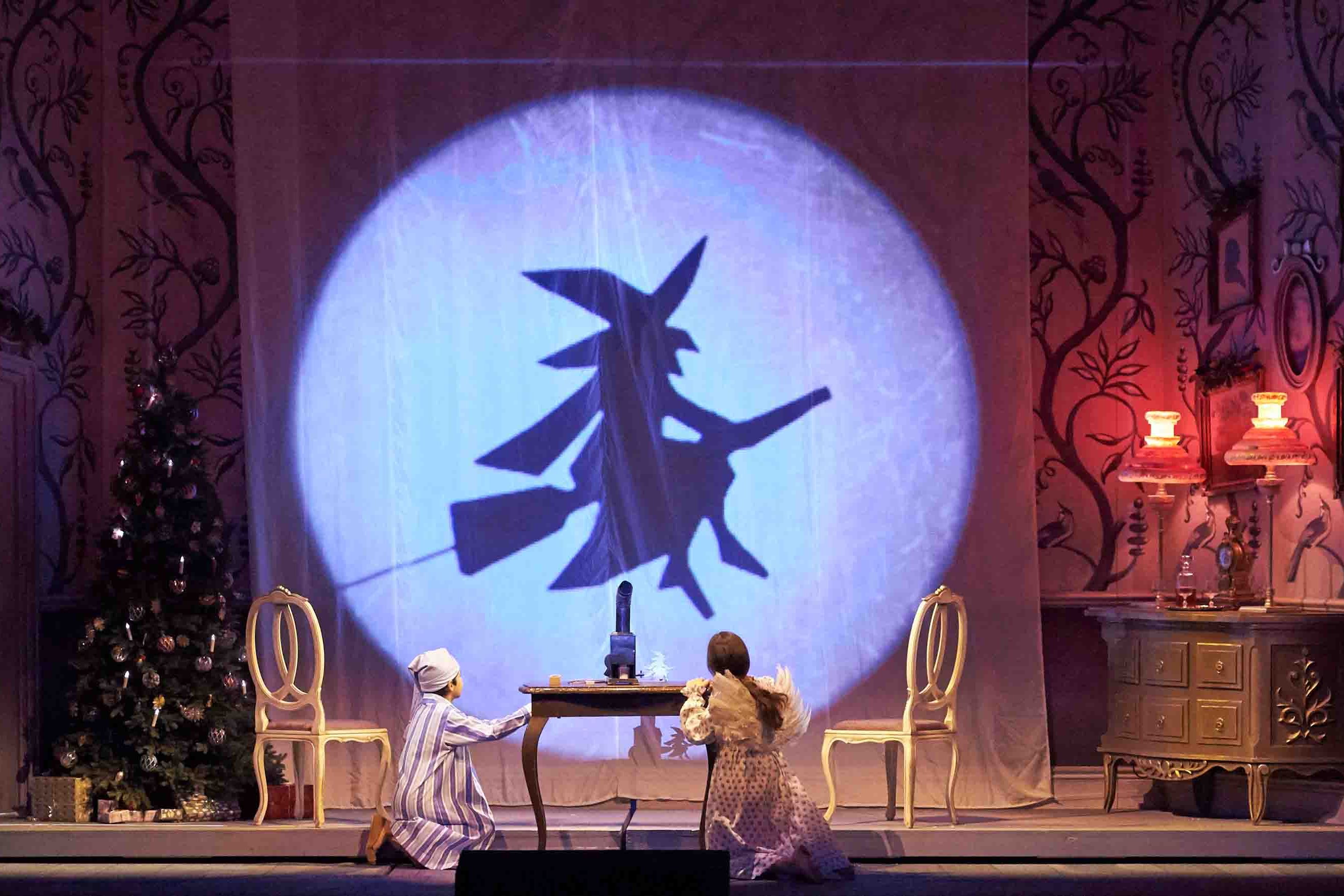 Wiener Staatsoper / Hänsel und Gretel @ Foto: Michael Poehn A scene from the family opera Hansel and Gretel. A boy and a girl in night dress kneel in front of a table in a historic children's room, a Christmas tree on the right, the silhouette of a witch on a broom in the background on a screen