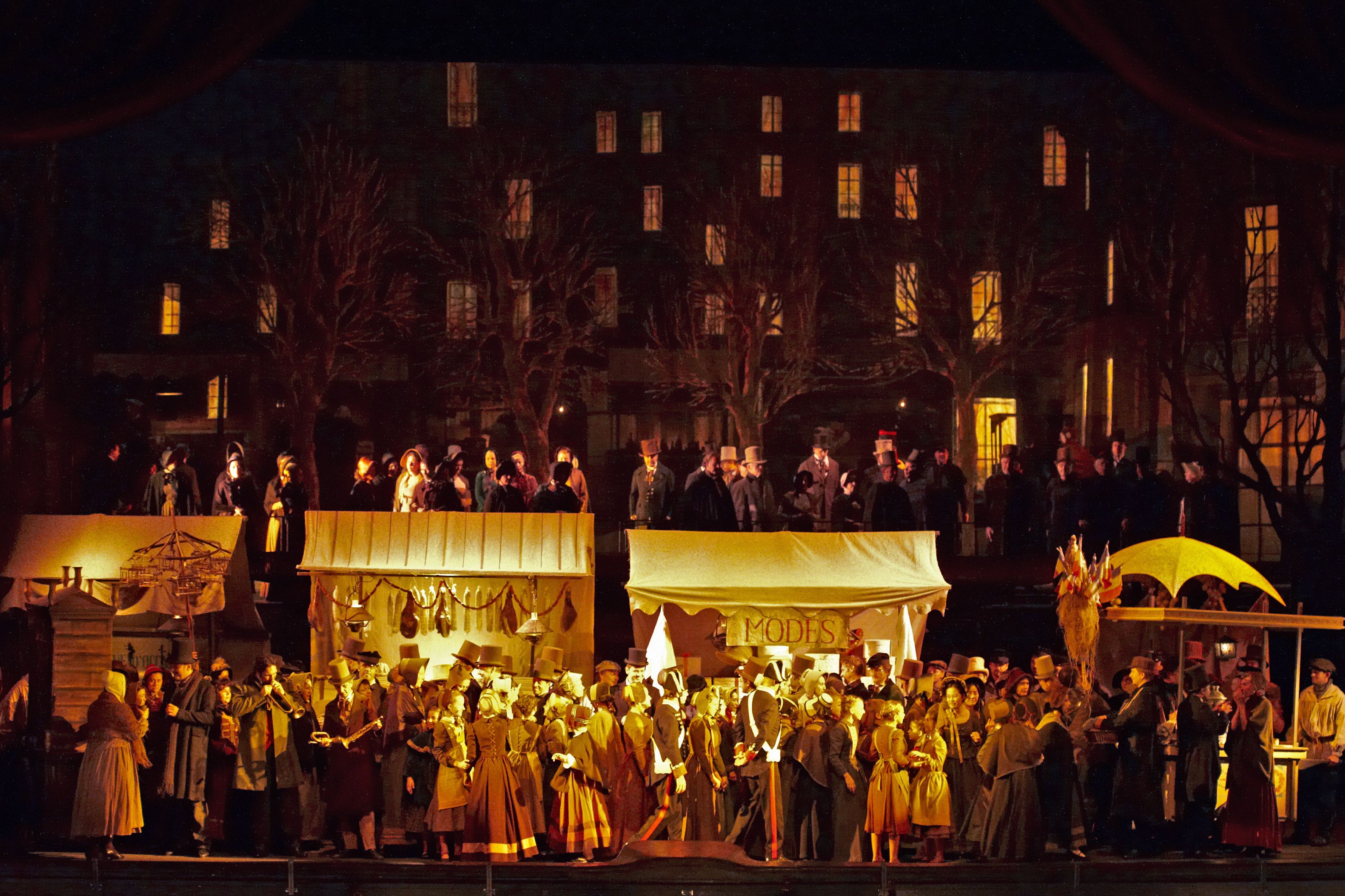 Wiener Staatsoper / La Boheme @ Foto: Michael Poehn Das Szenefoto aus der Oper "La Boheme" zeigt eine abendliche Szene auf einem Markt in gelbem Licht, vor den Ständen drängt sich eine große Menschenmenge in historischen Kostümen, im Hintergrund Bäume und hohe Häuser, deren Fenster erhellt sind