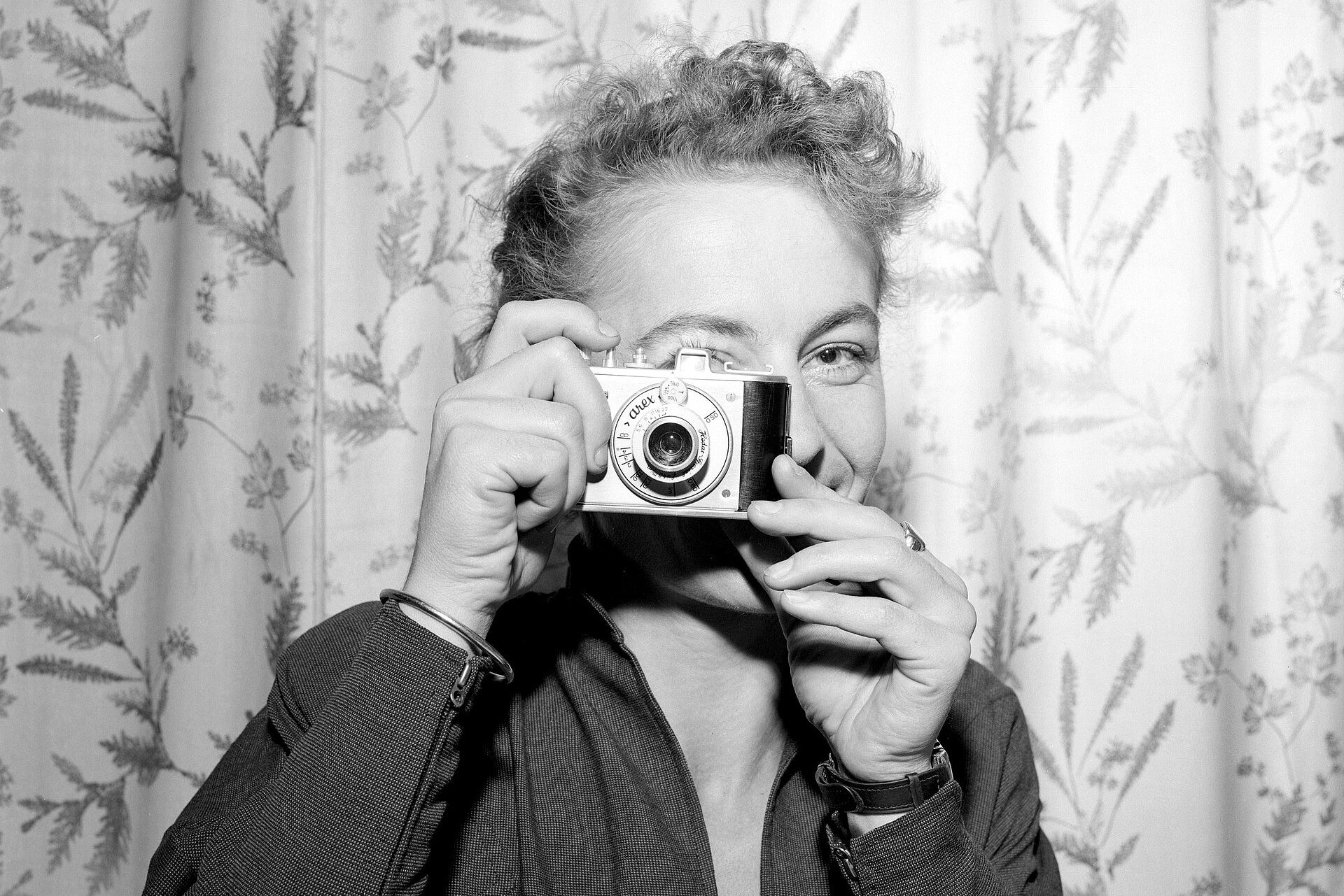 Das Schwarzweiß-Foto zeigt eine Frau, die sich eine historische Kamera vor ihr rechtes Auge hält und sich zum Betrachter / zur Betrachterin wendet wie wenn sie ein Foto machen würde